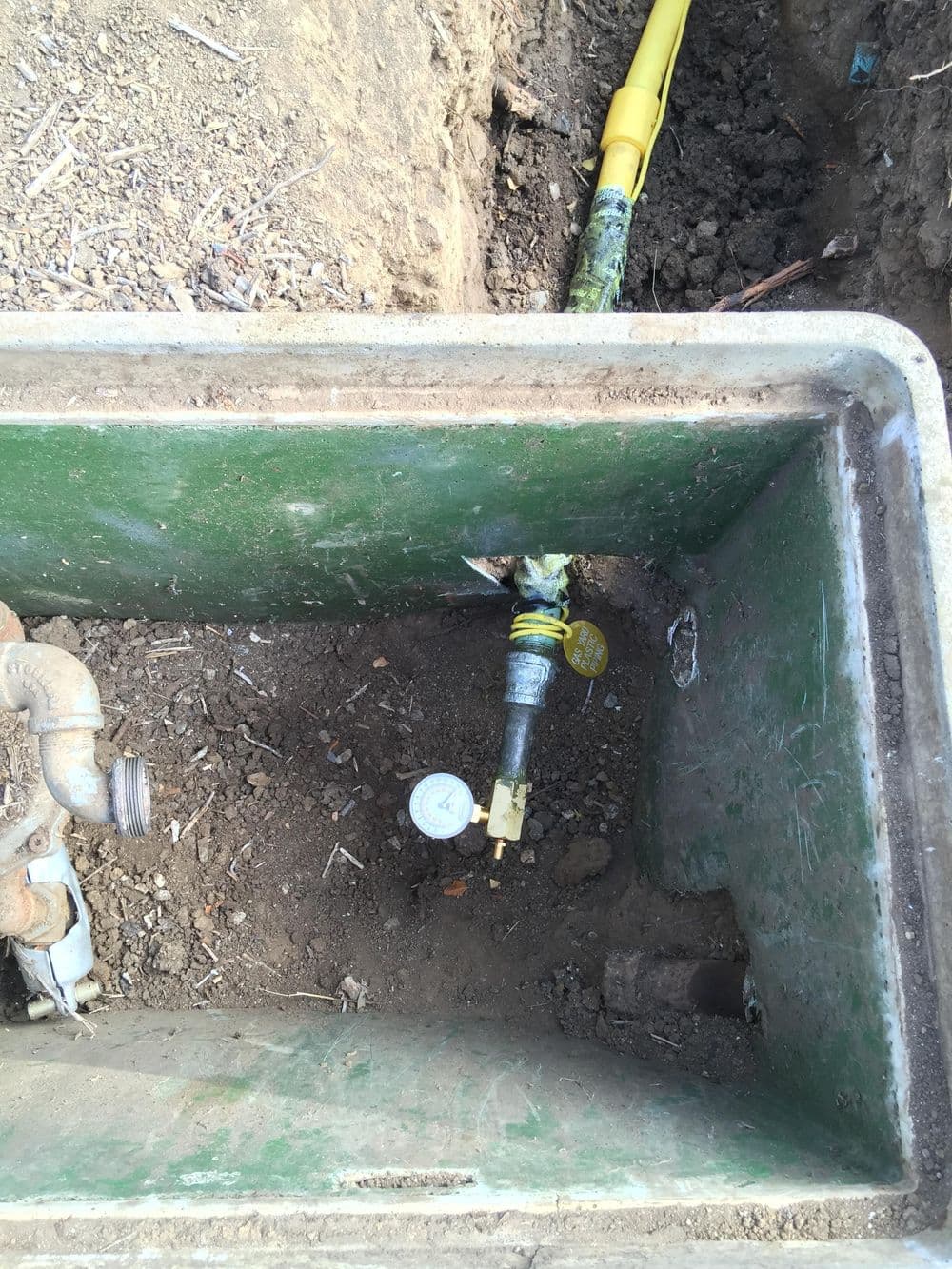 Water meter and valve inside a green utility box, showing soil and pipes.