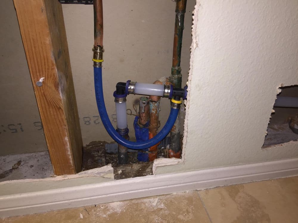Exposed plumbing with blue and clear pipes behind drywall, showing water connections.
