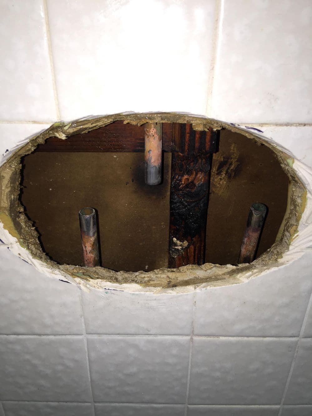 Hole in wall exposing plumbing pipes and wooden structure behind tiles. Home repair issue.
