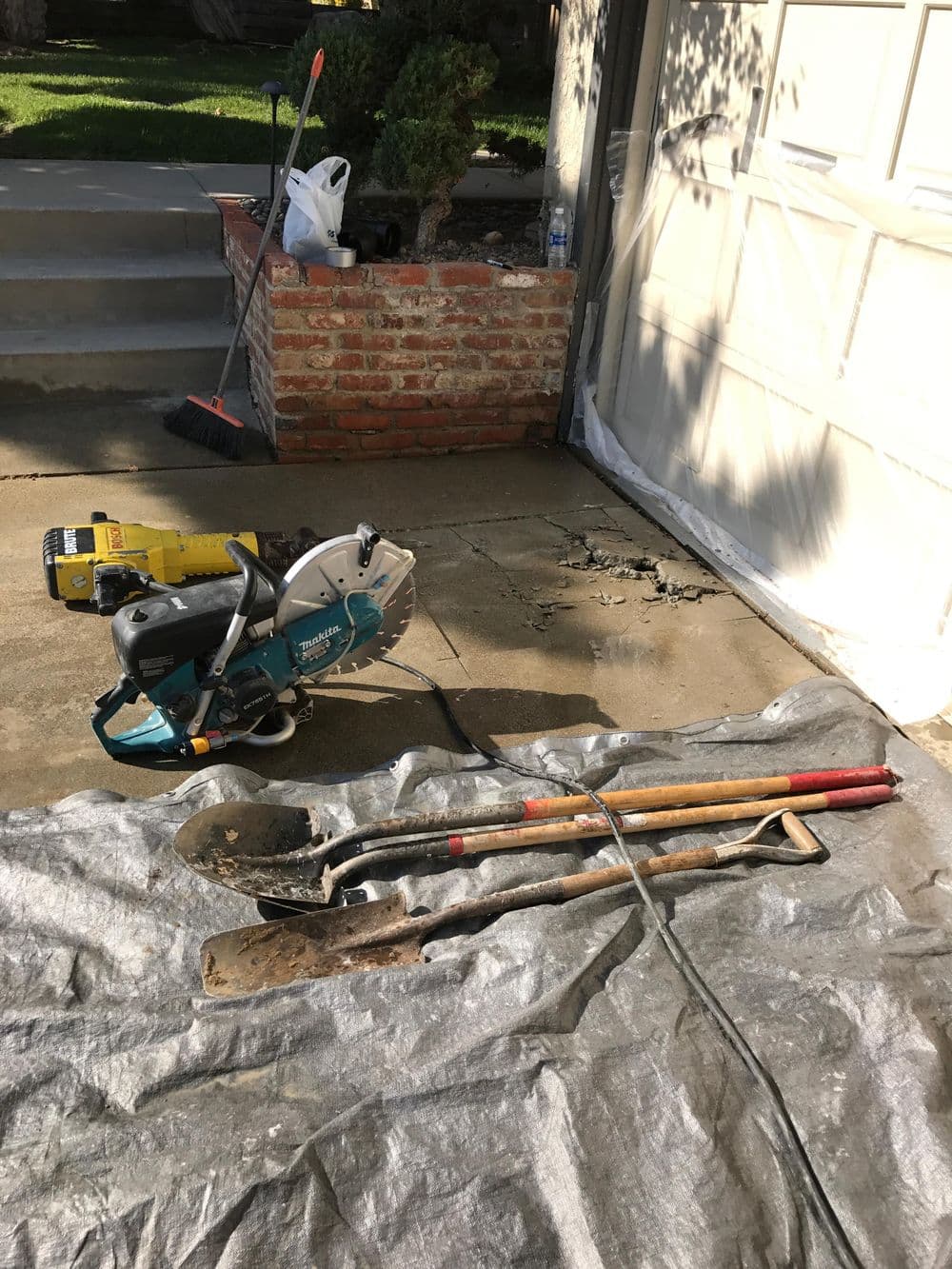 Power tools and shovels on a tarpaulin, with a residential garage and landscaping in the background.