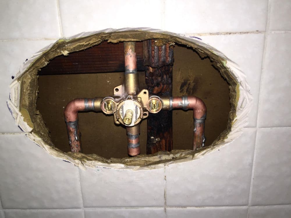Exposed plumbing pipes and valve behind a wall with tiled surface. Home repair context.