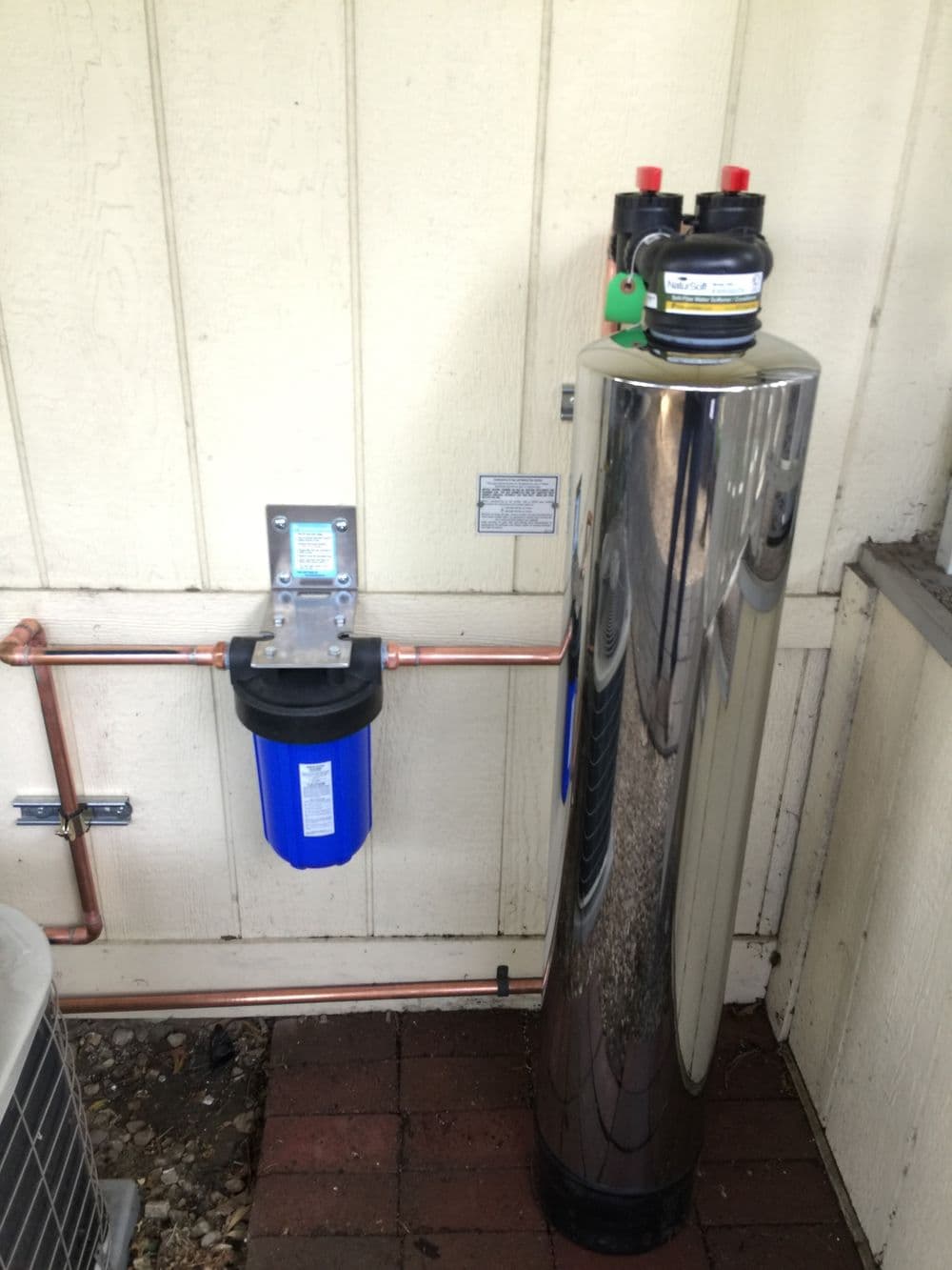 Stainless steel water filtration system with blue filter and copper piping installed outdoors.