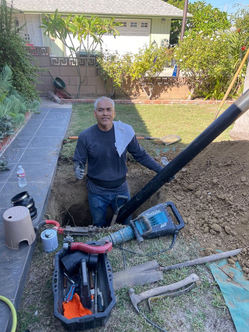 Man working in a garden excavation, holding a tool, with grass and garden plants visible.