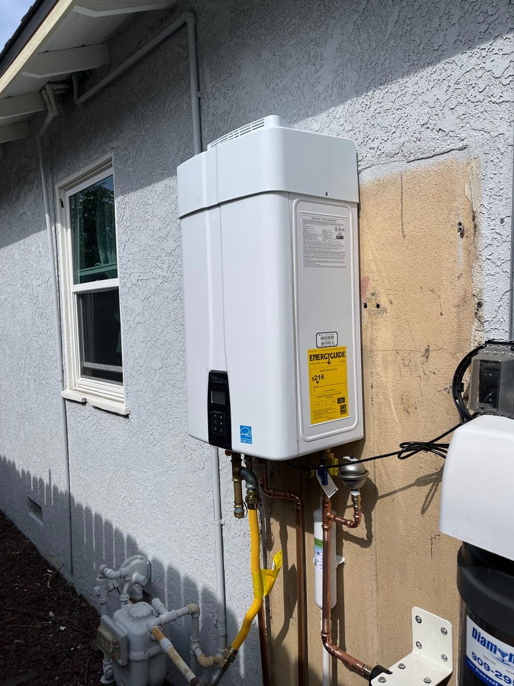 Tankless water heater installed on exterior wall of home, featuring energy guide label and pipes.