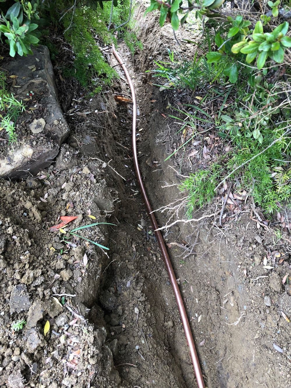 Copper piping laid in a trench surrounded by soil and vegetation.
