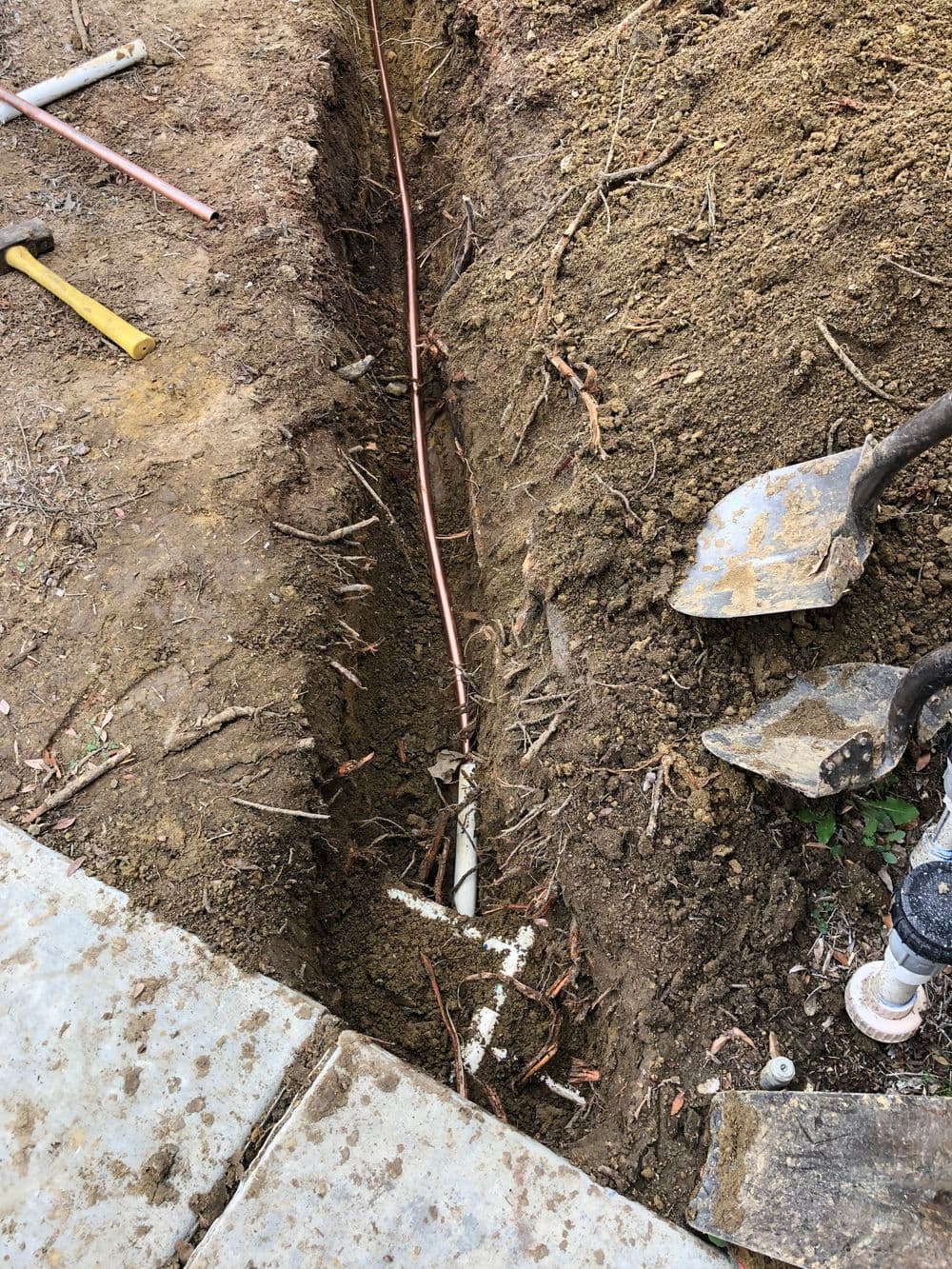 Excavated trench with copper piping and tools for plumbing installation outdoors.