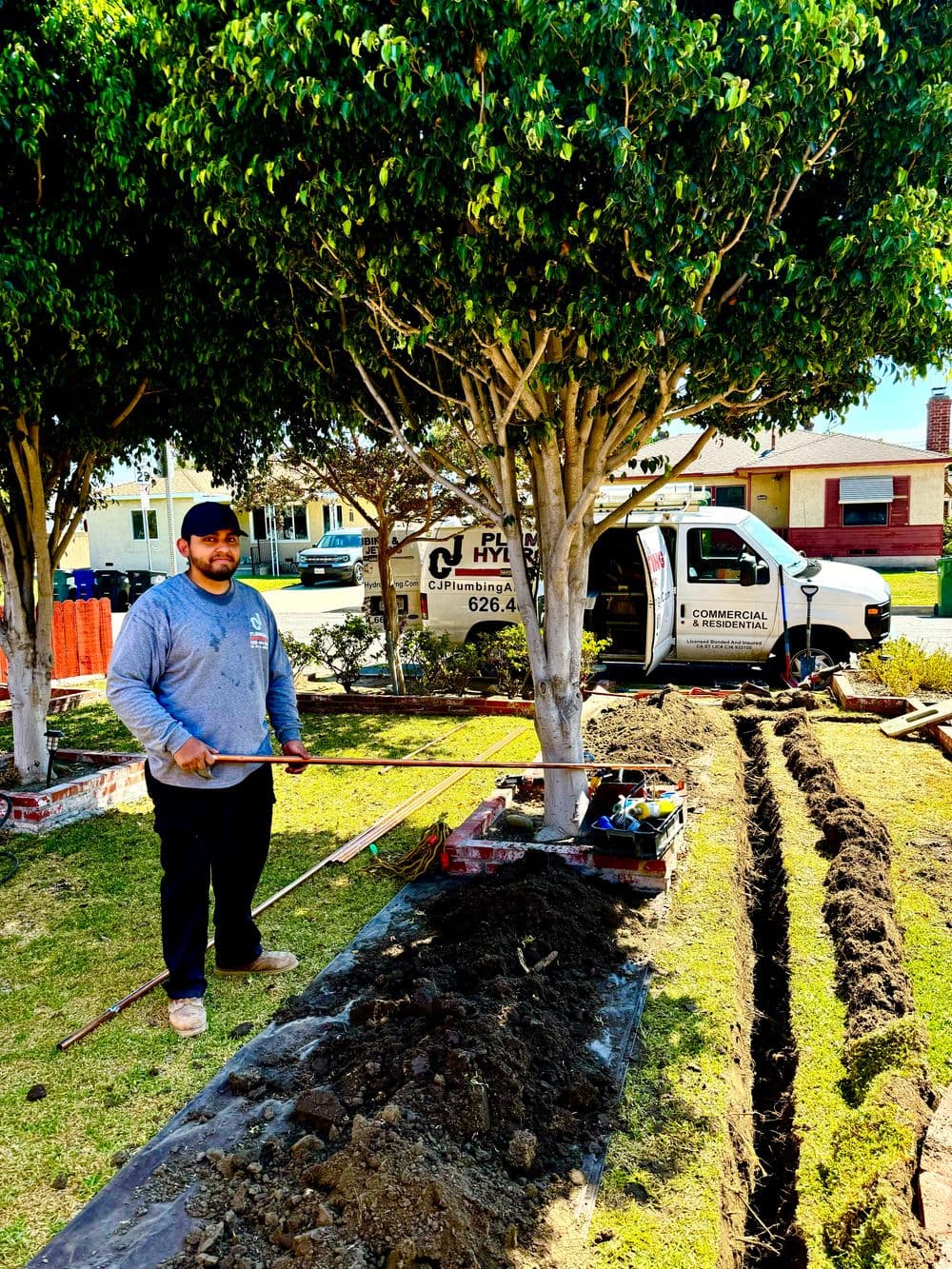 Landscaper working with tools beside trees and a plumbing truck on a residential lawn.