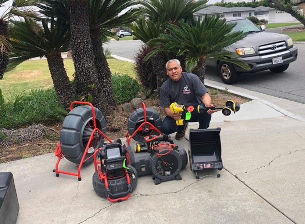 Man holding plumbing tools next to drainage equipment on a driveway with palm trees.
