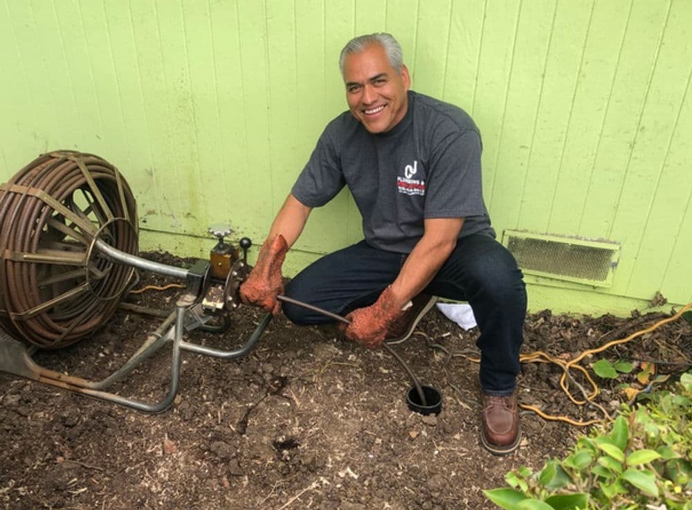 Man using a plumbing snake to clear a drain outside a green wall. Tools and dirt visible.