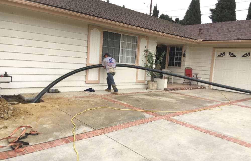 Worker installing a large black pipe outside a residential home with a concrete driveway.