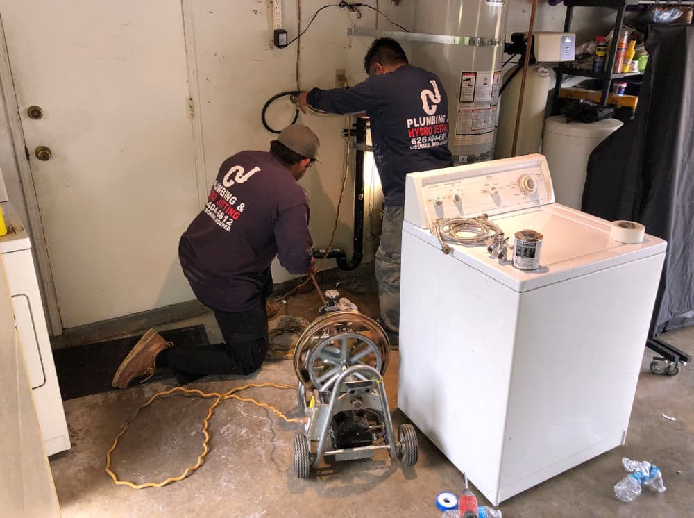 Plumbers repairing a pipe with a drain cleaning machine in a garage setting.